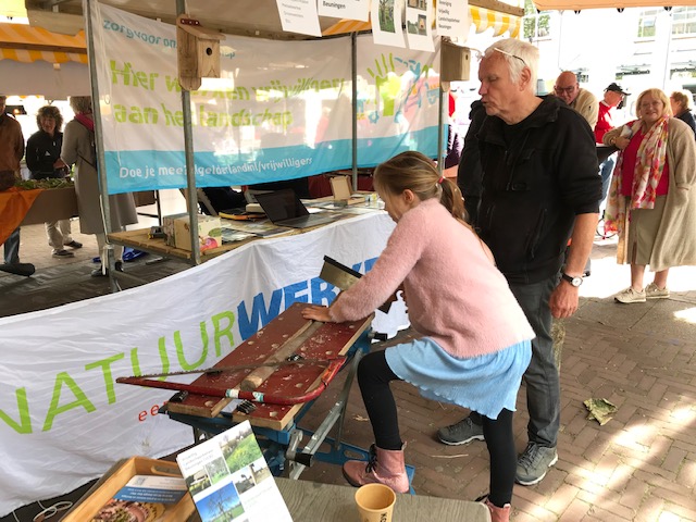 Lotte zaagt een tak door. Lotte met roze vest en paardenstaart, één voet op het onderstel van de werkbank.