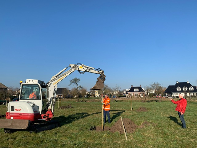 Op een grasveld staat helemaal links een graafmachine, In het midden en rechts twee mannen in oranje jassen
