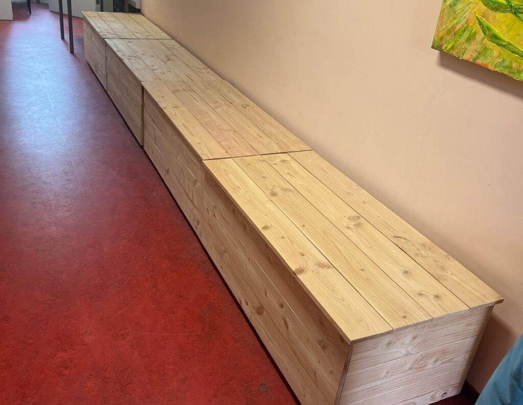 Vier blankhouten kisten (met deksels) naast elkaar vormen samen een lange zitbank.